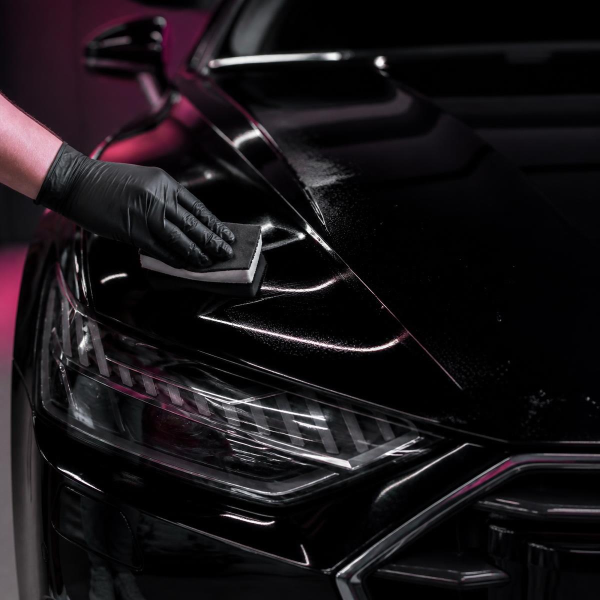 Gloved technician spreading AG Formula 1051 paint protection coating across the glossy black hood of a luxury car