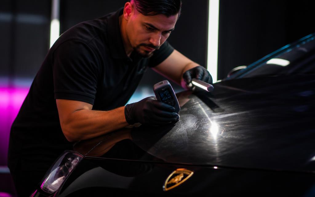 Technician measuring paint depth and inspecting swirl marks on the black hood of an exotic supercar